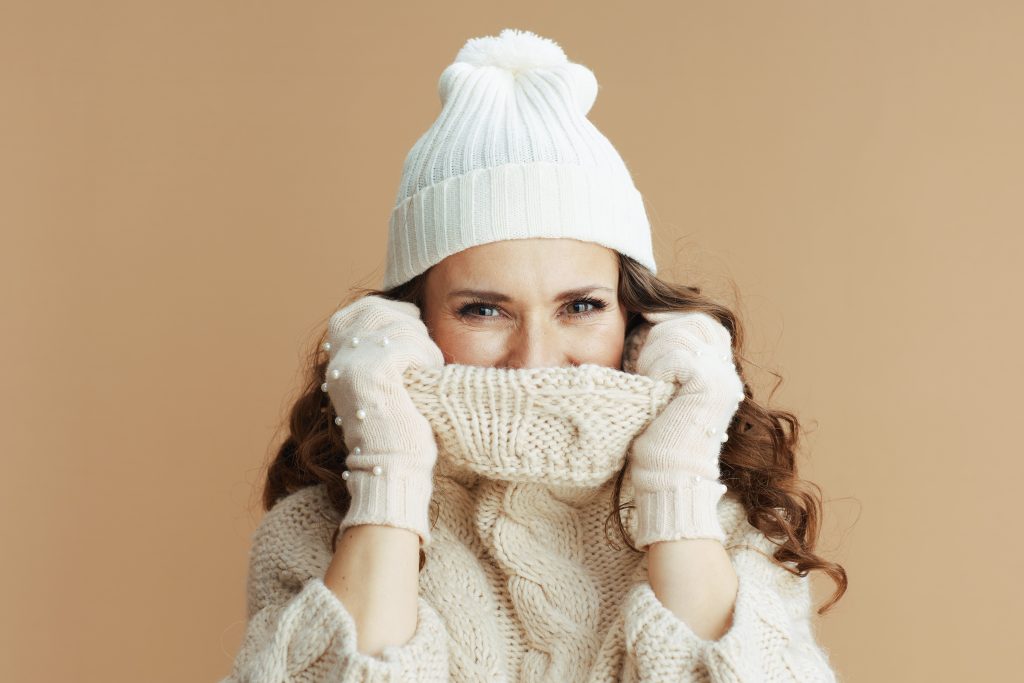stylish middle aged woman in beige alpaca sweater, mittens and hat wrapped in a collar isolated on beige